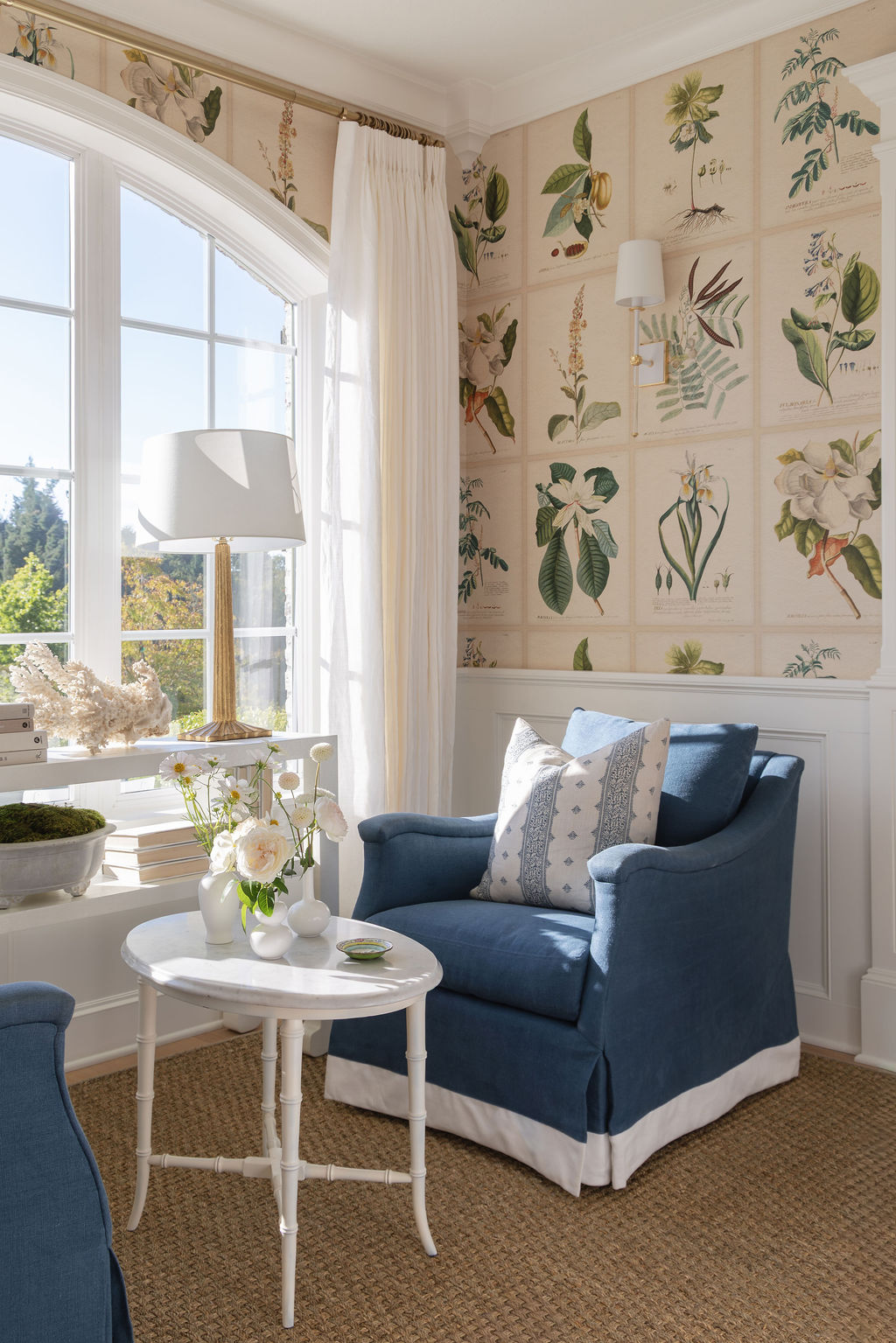 Sunny sitting room with two blue skirted chairs, white bamboo side table, white lamps, and vintage botanical wallpaper around arched windows.
