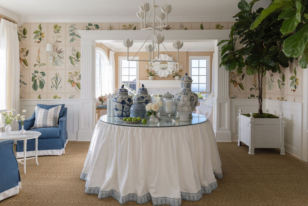 Centered view of the round skirted table styled with blue-and-white ginger jars, looking toward a living room framed by columns and botanical wallpaper.