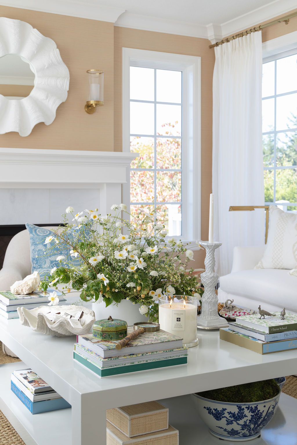 Coffee-table close-up with a loose, garden-style arrangement of white flowers, books, a shell bowl, candlesticks, and a lit candle in a bright white living room.