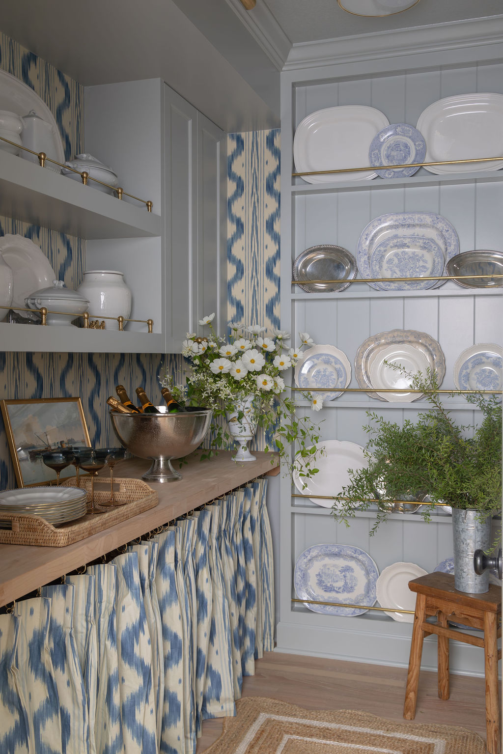 Butler’s pantry with blue ikat skirted base, pale gray shelves with brass rails, vintage silver, white china, and wildflowers in a vase.