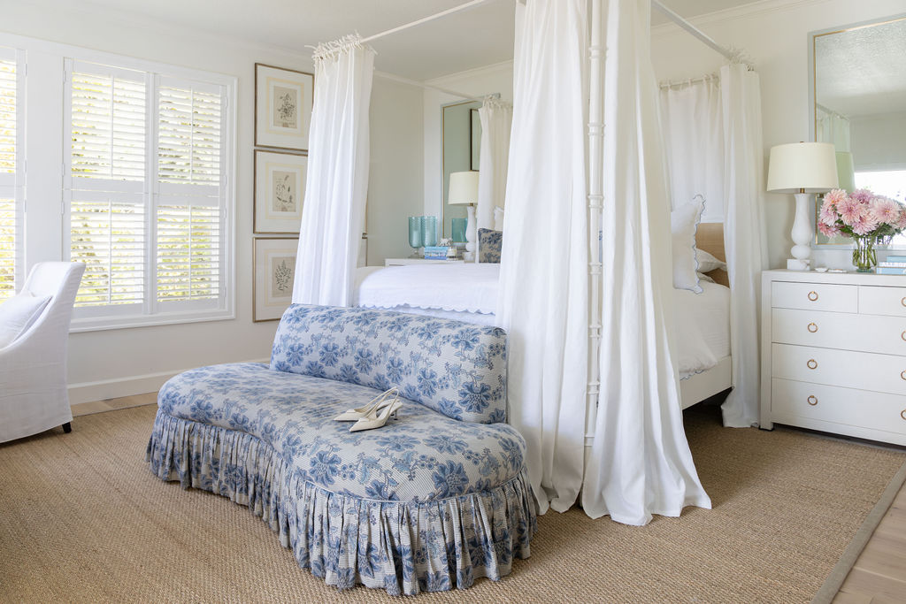 Primary bedroom with white four-poster canopy bed, seagrass rug, and a blue floral curved bench with ruffled skirt at the foot.