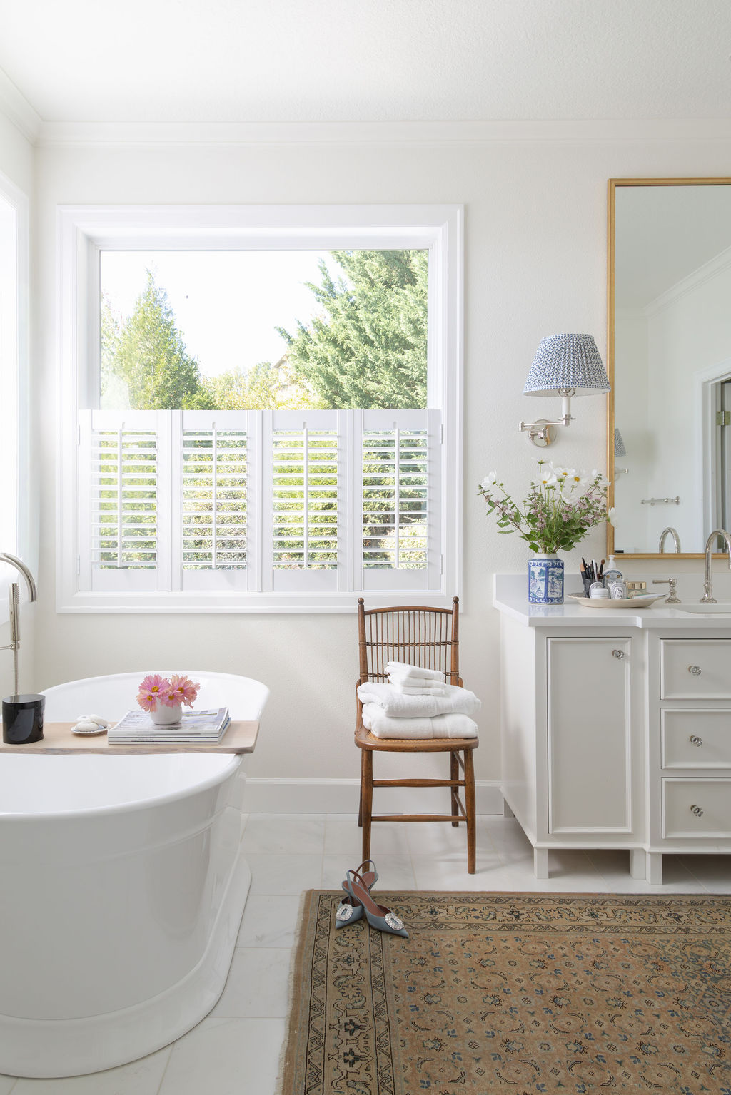 Bathroom detail: tub by a window with café shutters, small wood chair stacked with towels, blue sconce, and fresh flowers on the counter.