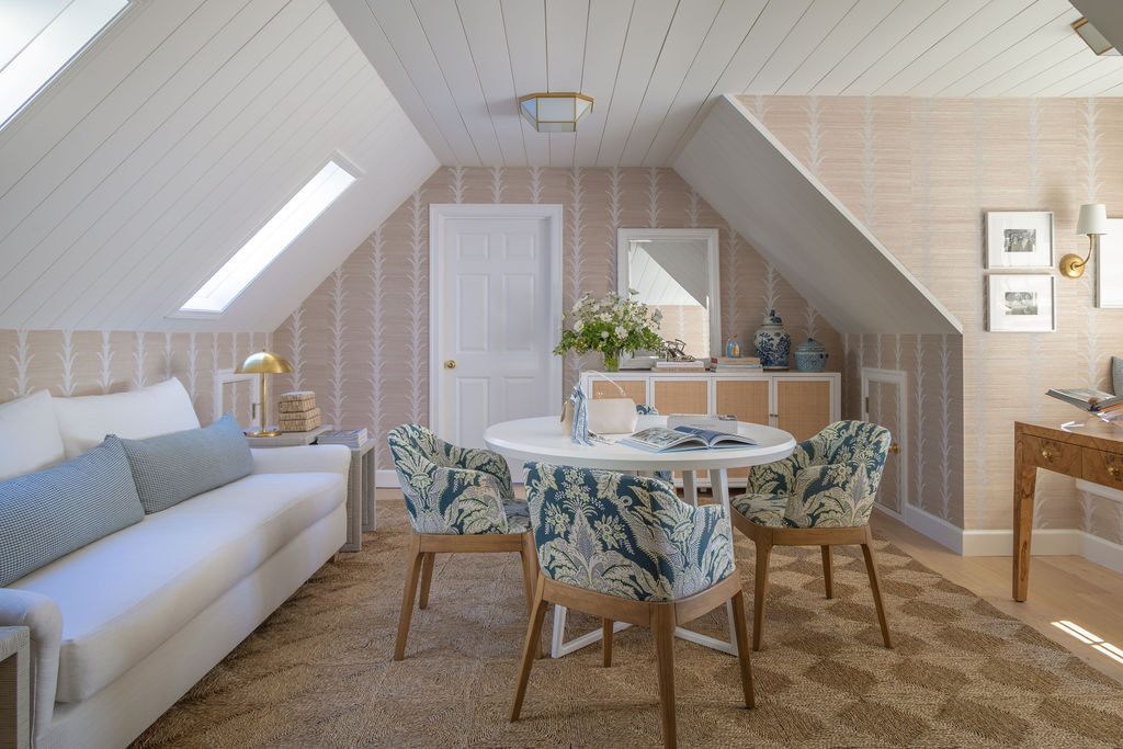 Attic studio with sloped ceilings, neutral grasscloth walls, white sofa, round white table, and four blue botanical upholstered chairs.