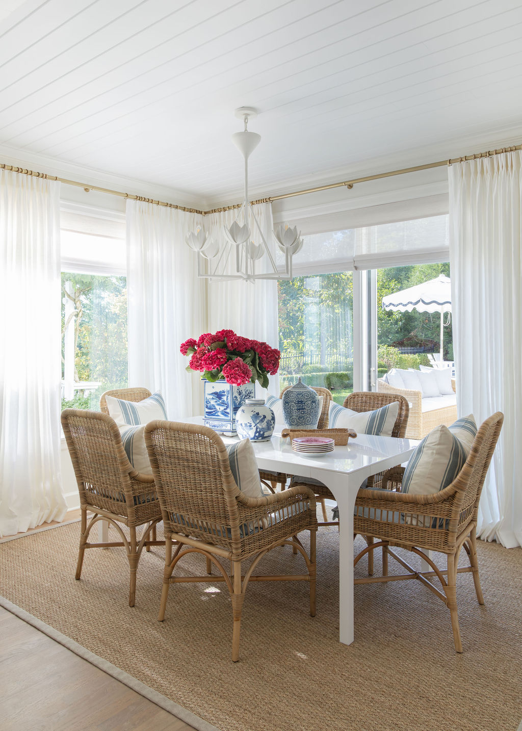 Sunny dining area with white table, six wicker armchairs with striped pillows, sheer white drapery, and white lotus chandelier.