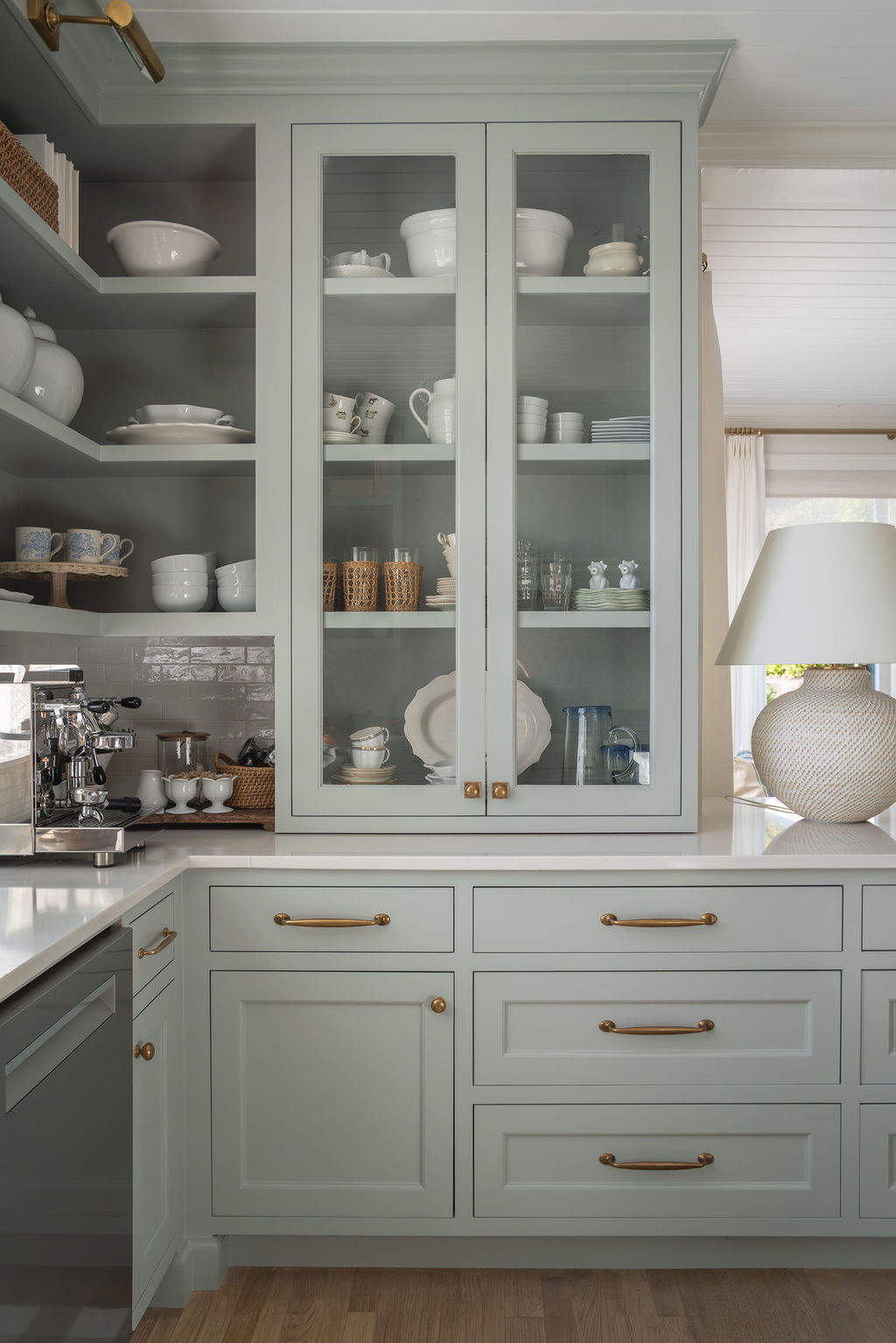 Sage green pantry cabinets with glass doors displaying white dishes and glassware, brass hardware, and a coffee station on white counters.
