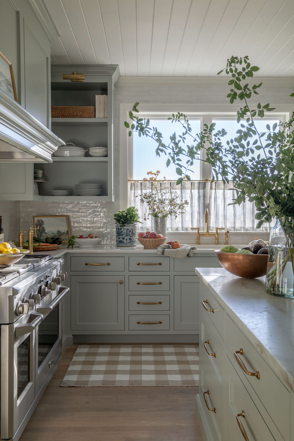 Kitchen range wall and island: gray-green cabinetry, brass hardware, copper bowl and greenery on the island, café curtains over the sink, and a beige-and-white checked rug on wood floors.