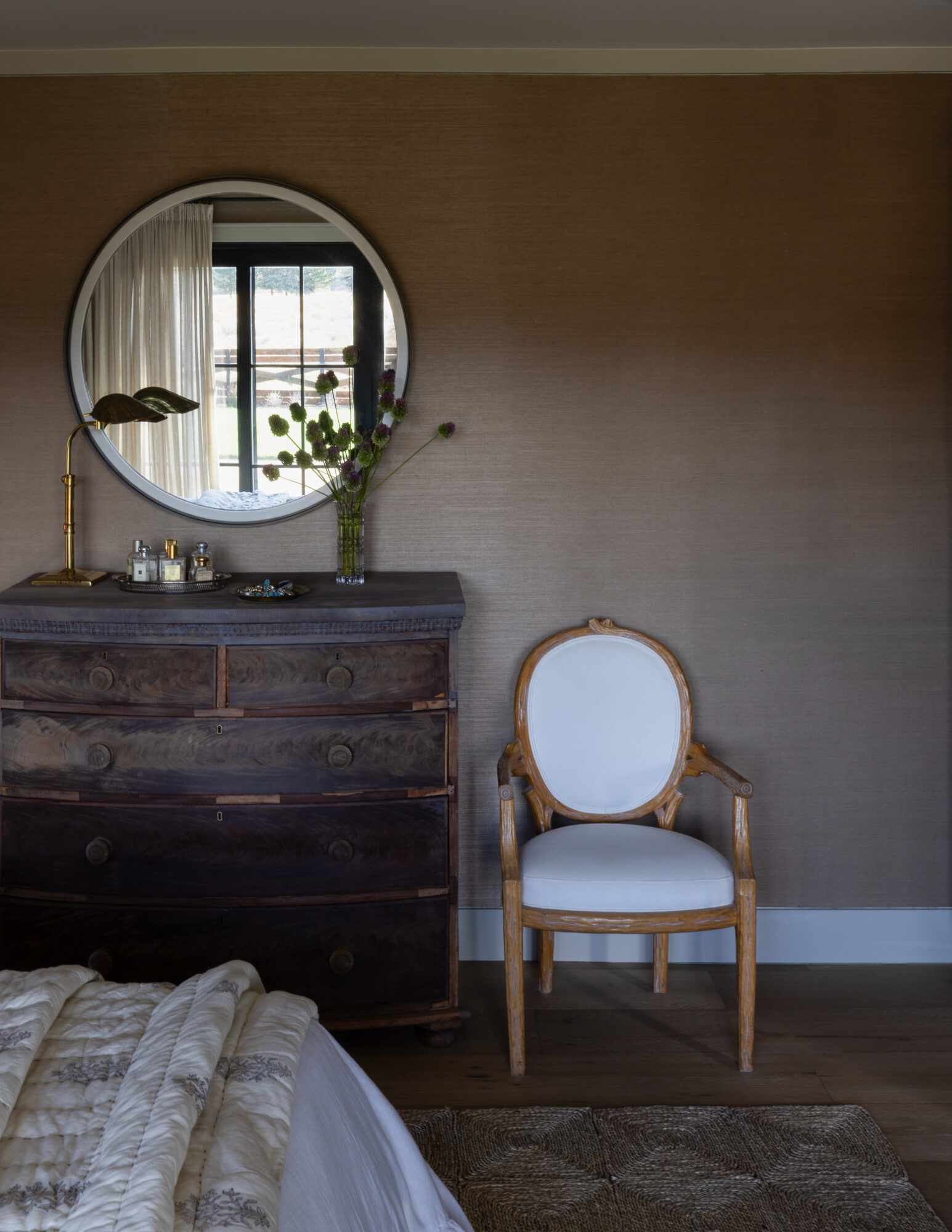 A serene bedroom corner featuring a vintage wooden dresser with ornate details, styled with a brass leaf table lamp, a round mirror reflecting natural light, and a vase of thistle blooms. A French-inspired wooden armchair with white upholstery sits beside the dresser, resting on a natural woven rug, creating a tranquil and timeless aesthetic.