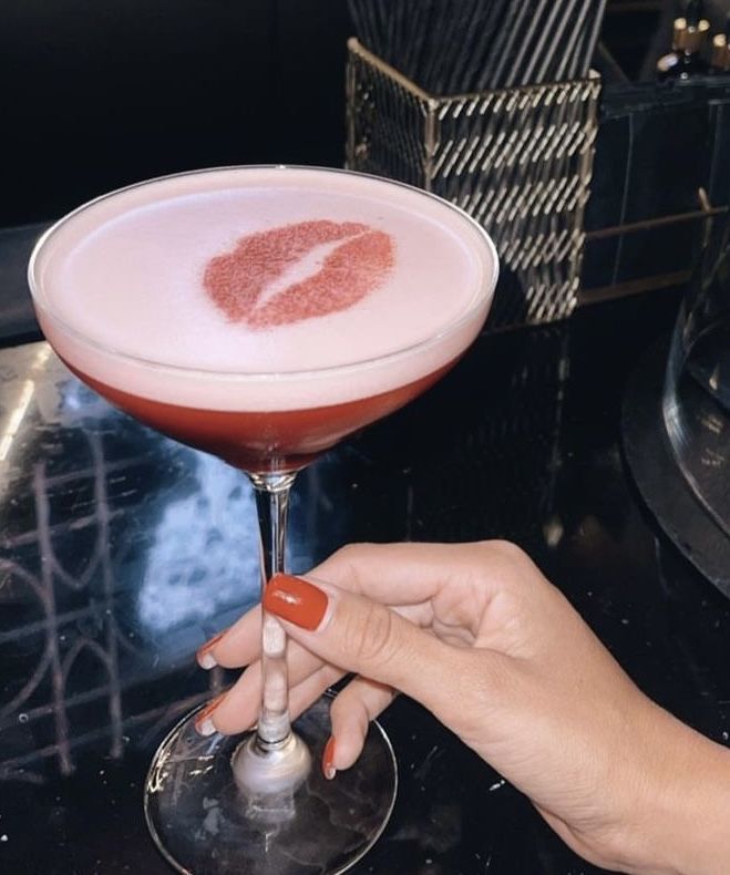 A chic Valentine's Day cocktail featuring a classic Cosmopolitan with a frothy egg white top, adorned with a lip stencil design in beet powder, held by a hand with red-painted nails against a stylish black bar background.