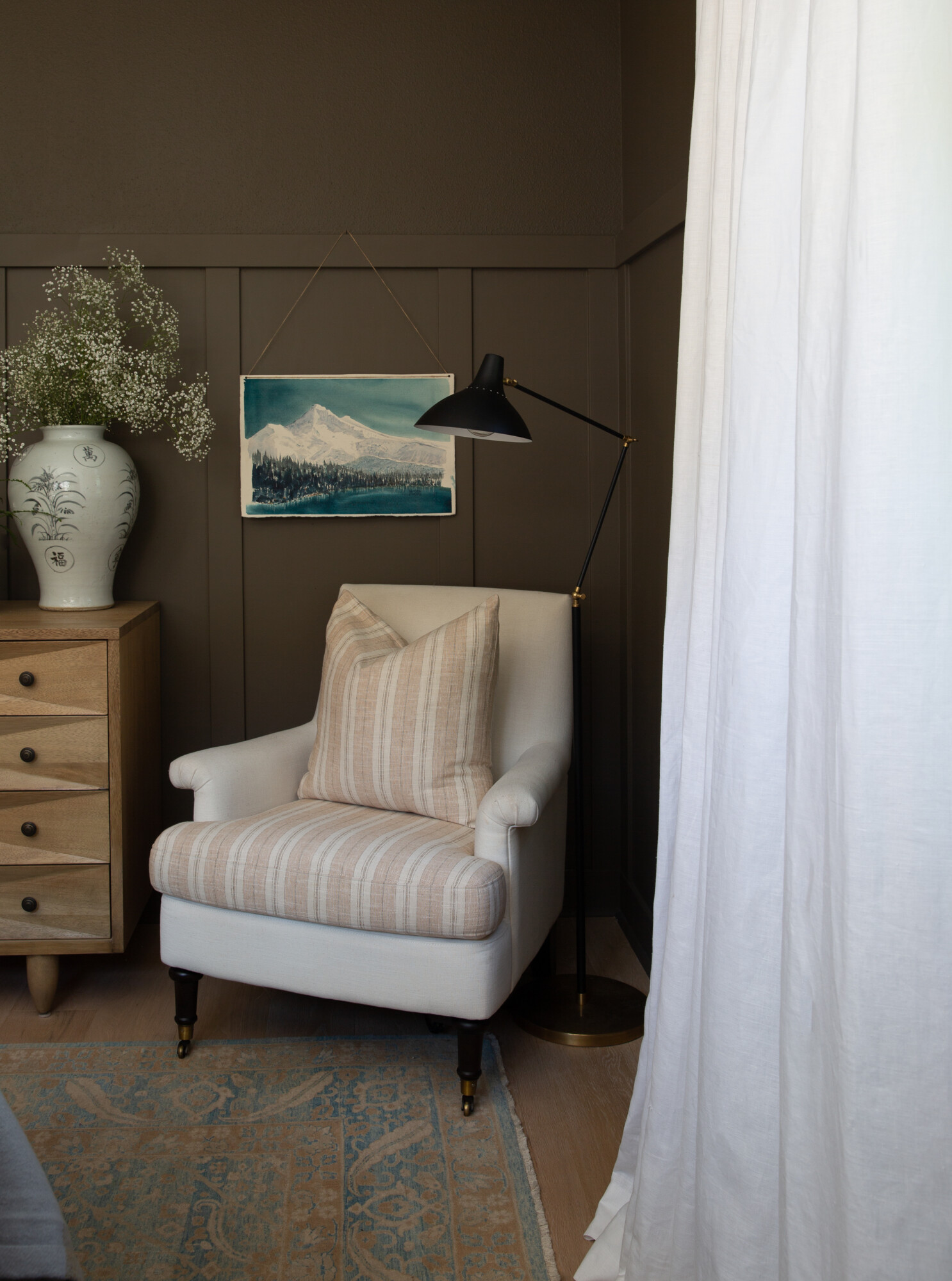 Cozy reading nook with a cream armchair featuring striped upholstery and a matching pillow, set against a deep olive paneled wall. A black and brass floor lamp provides lighting beside a light wood chest of drawers with a ceramic vase and baby's breath. A blue and tan vintage rug adds warmth to the wood floor, and white linen curtains softly frame the space.