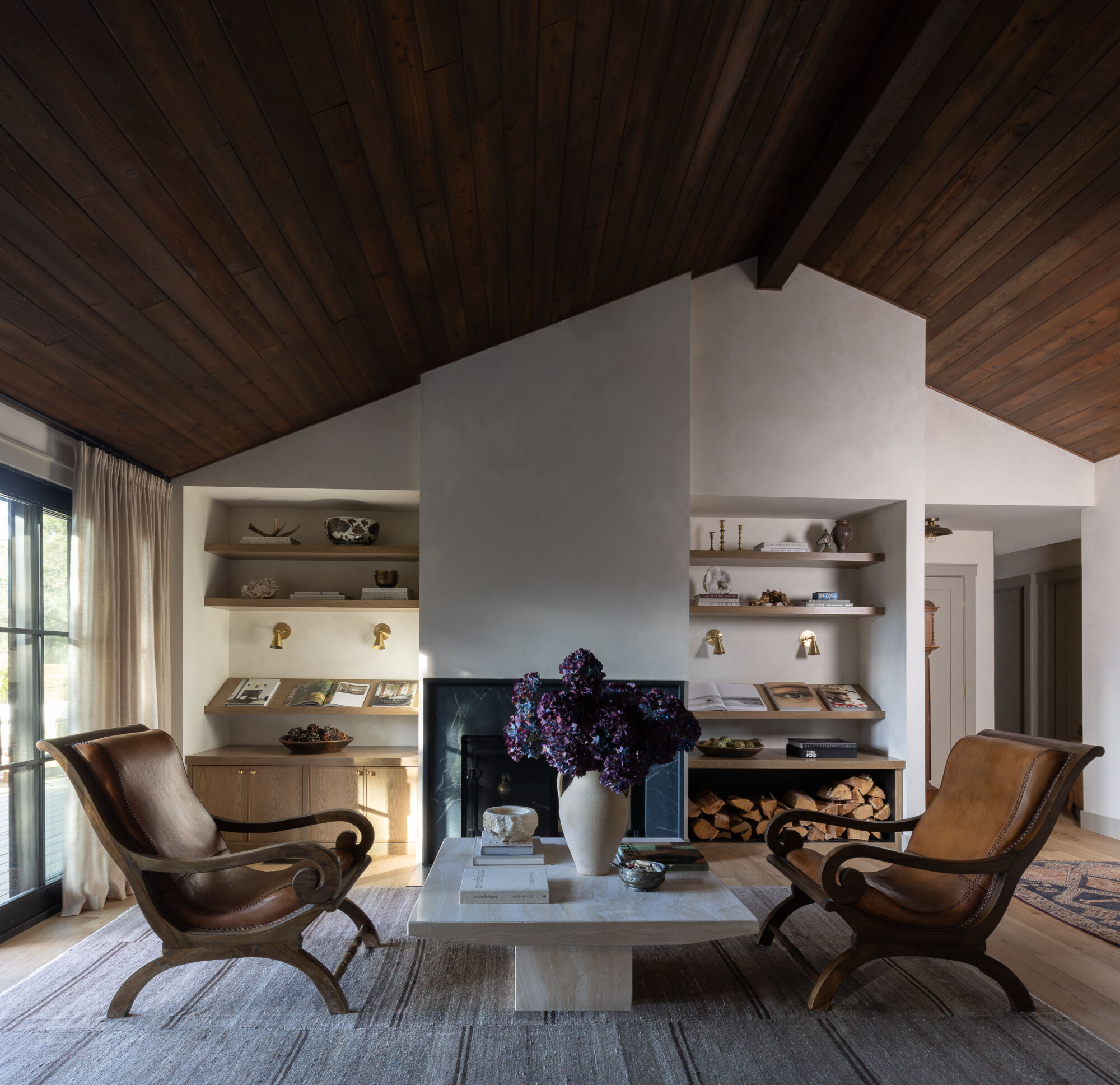 Warm and inviting living room featuring two sculptural leather accent chairs with curved wood frames facing a marble coffee table topped with a large vase of deep purple flowers. Built-in shelves flank a minimalist plaster fireplace, styled with books, objects, and brass picture lights. A dark wood plank ceiling adds depth and contrast to the soft, neutral-toned walls and natural wood floors.