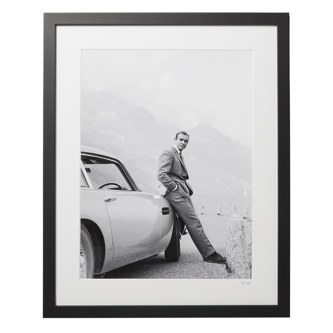 Framed black and white photo – A stylish black and white photograph of a man in a suit leaning against a vintage car with mountains in the background, framed in black.
