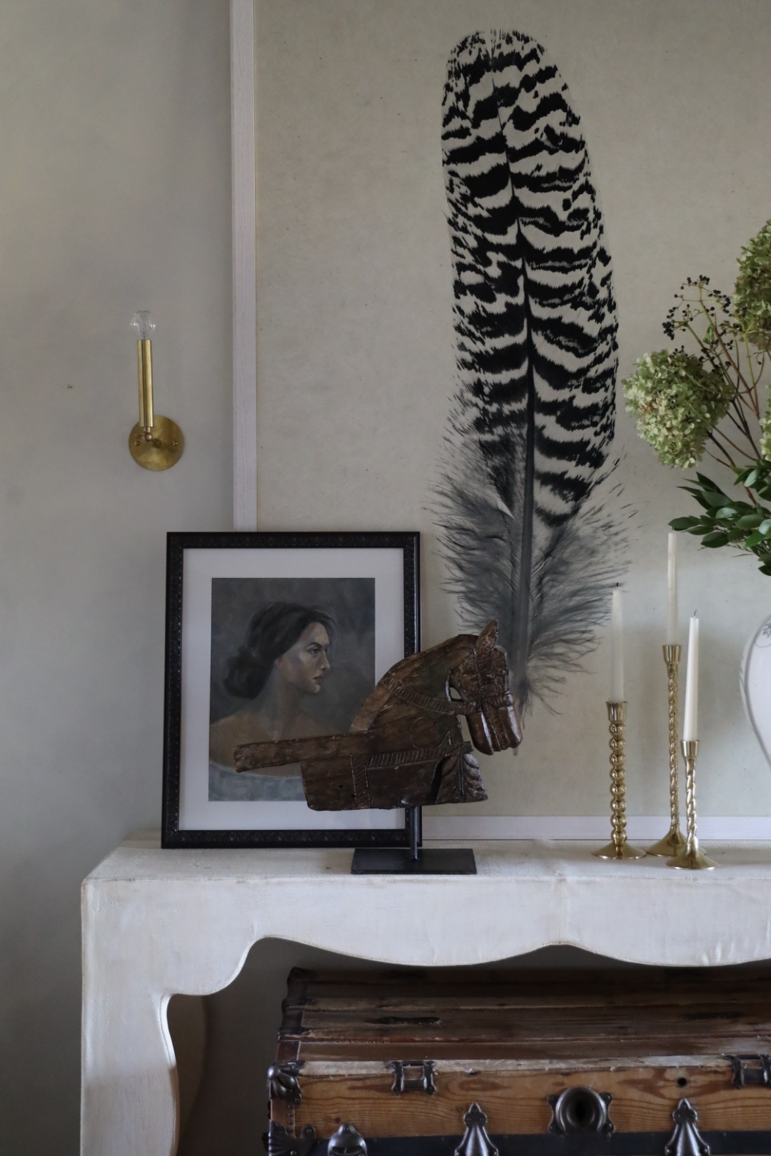 A layered and eclectic vignette featuring a large feather print, a vintage-style portrait of a woman in a black frame, a carved wooden horse sculpture on a stand, and a mix of gold-toned twisted candlesticks. The arrangement sits on a white scalloped-edge console table, with an antique wooden trunk placed beneath it and a brass wall sconce to the side. Hydrangeas and greenery peek in from a vase on the right, adding a fresh, organic touch.