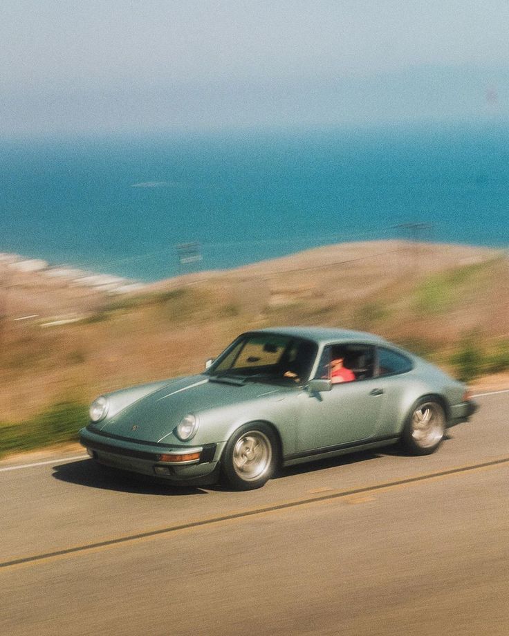 Vintage Porsche driving along a coastal highway with the ocean in the background, captured in a blurred motion style that evokes a nostalgic, cinematic feel.