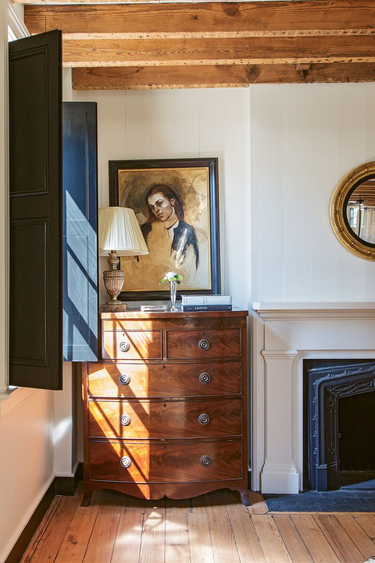 sunlit image of a corner of a room with shuttered windows open and and antique chest decorated with vintage artwork and a lamp