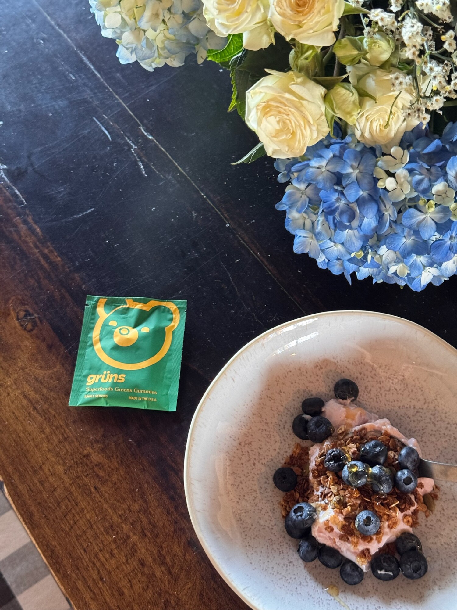 A packet of Grüns Superfoods Greens Gummies sits beside a breakfast bowl topped with granola, blueberries, and honey, with a lush bouquet of white and blue flowers in the background—an easy, nourishing morning moment.