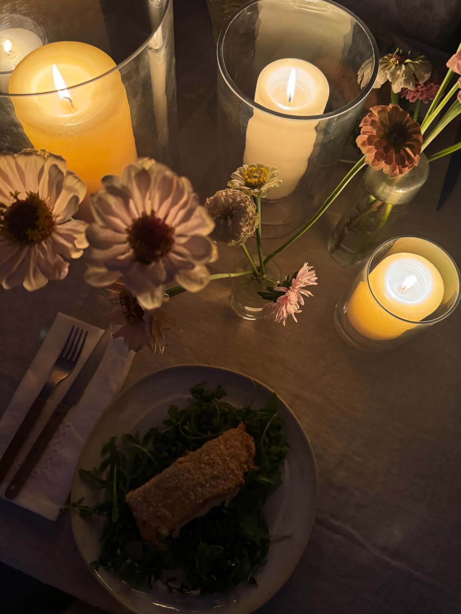 A romantic evening tablescape lit by glowing hurricane candles and soft pink zinnias in glass bud vases, with a plate of crispy stuffed chicken over arugula and vintage-style flatware—capturing the cozy magic of dinner under the stars.
