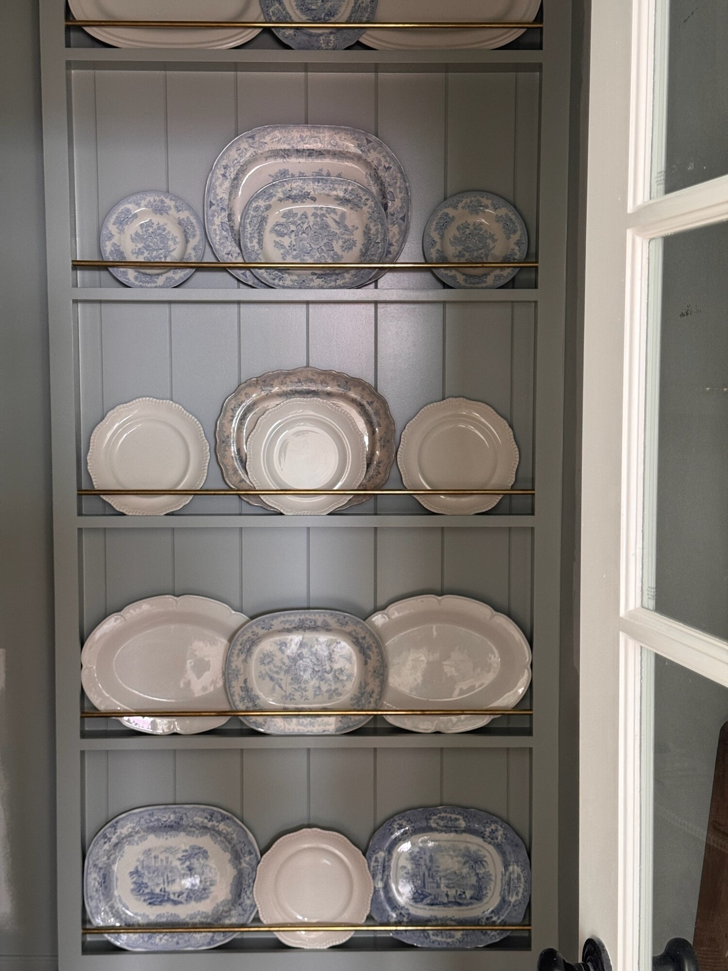 Close-up view of a built-in plate rack painted in soft blue, featuring an artful display of vintage white and blue transferware platters and dishes. Brass rails hold each shelf in place, adding a touch of elegance and shine. The curated collection creates a charming and timeless focal point within the pantry.