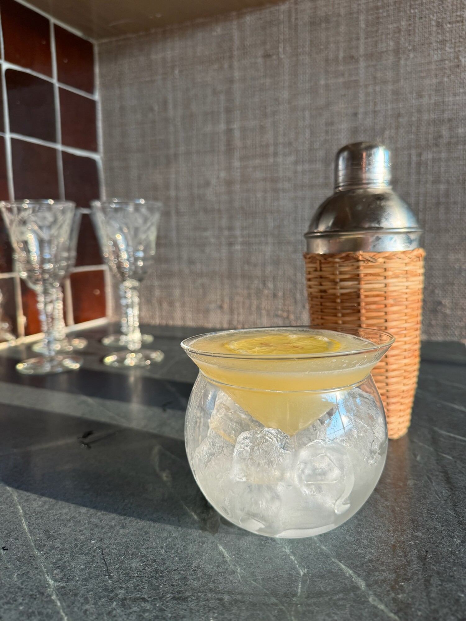 A citrus cocktail garnished with a lemon wheel served in a double-walled chiller glass over ice, set on a dark stone countertop with a vintage wicker-wrapped cocktail shaker and crystal stemware in the background—elevated summer sipping at golden hour.