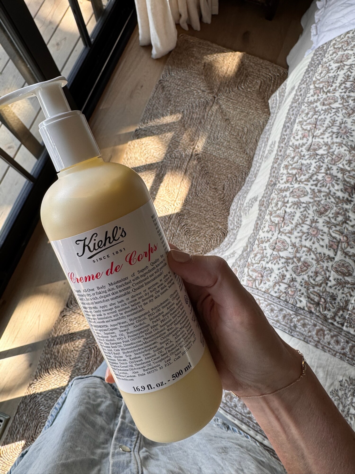 A hand holding a bottle of Kiehl’s Creme de Corps body moisturizer in a cozy bedroom setting, sunlight streaming through the window—hydration made chic.