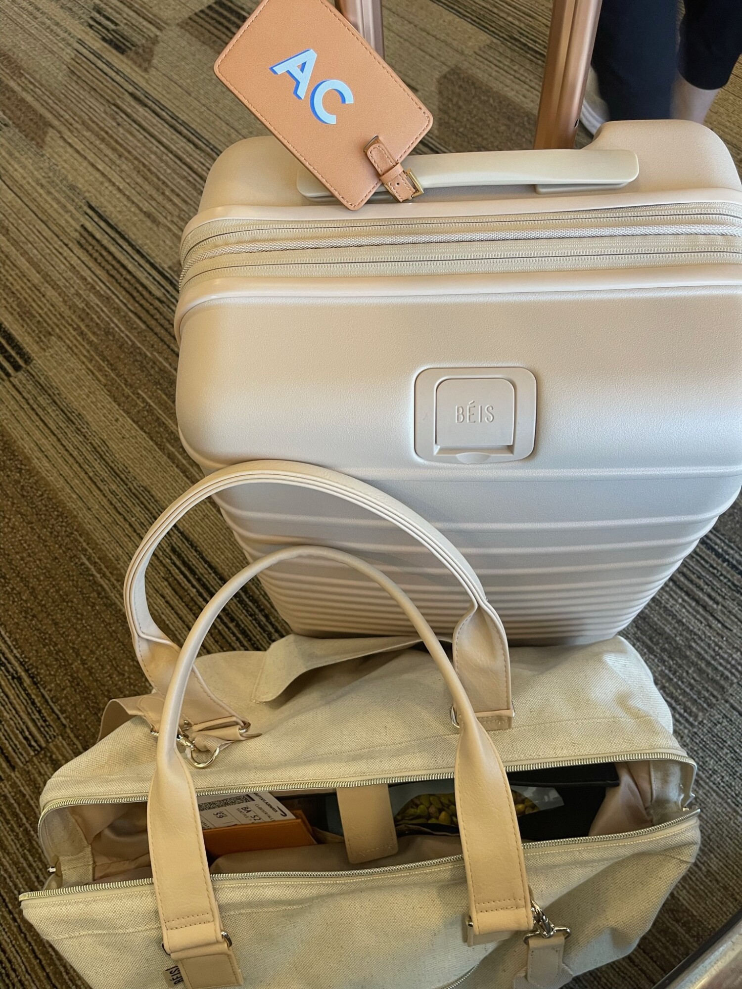 A beige Beis carry-on and weekender bag with a monogrammed tan luggage tag sit ready for travel on an airport floor—organized and stylish on-the-go essentials.