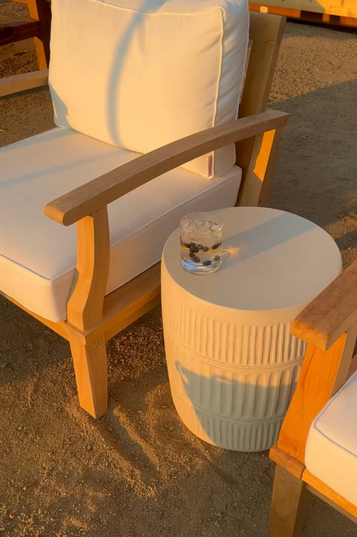 Two wood-frame outdoor lounge chairs with white cushions sit beside a textured ceramic side table holding a chilled drink with berries, all bathed in the soft glow of golden hour—simple and serene backyard perfection.