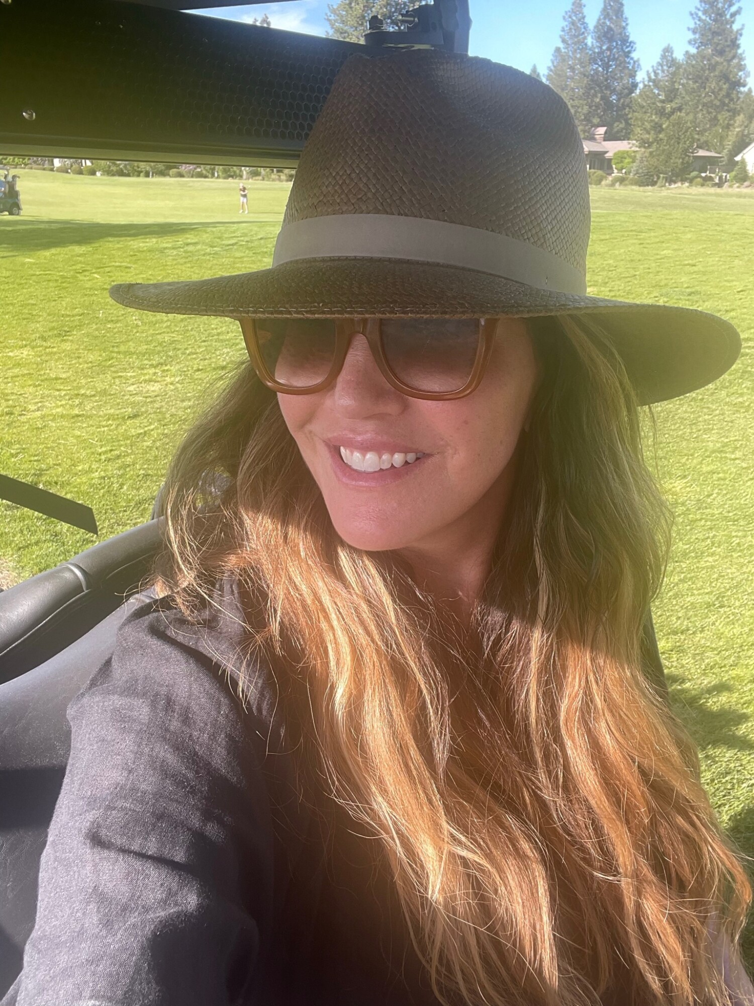 A sunlit selfie of a woman in oversized sunglasses and a structured straw hat, smiling in a golf cart on a bright summer day—classic sun-safe glam.