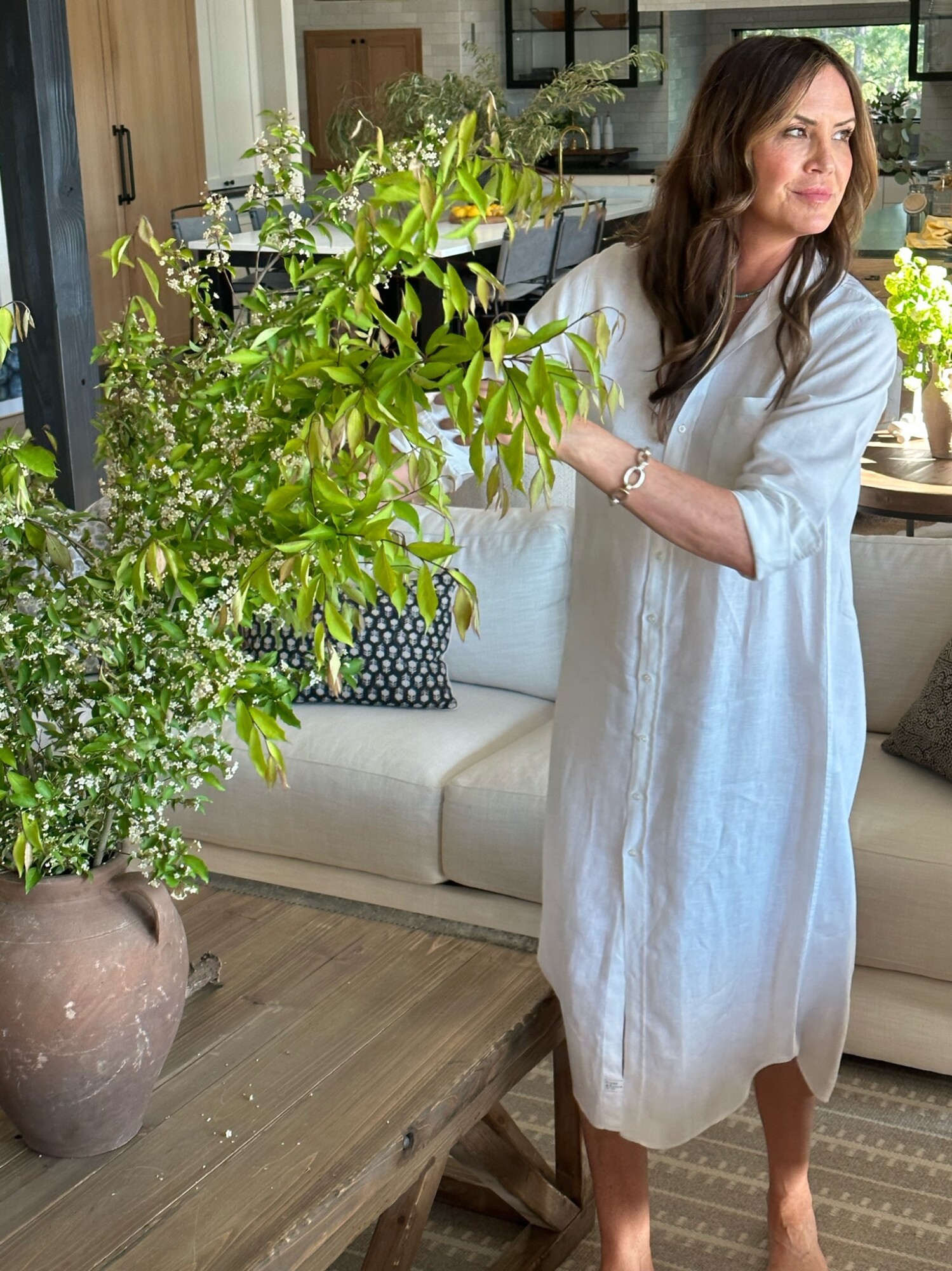 A woman styling fresh greenery in a rustic vase while wearing a long, white linen button-up dress in a warm, inviting living room—easy summer style meets effortless hosting.