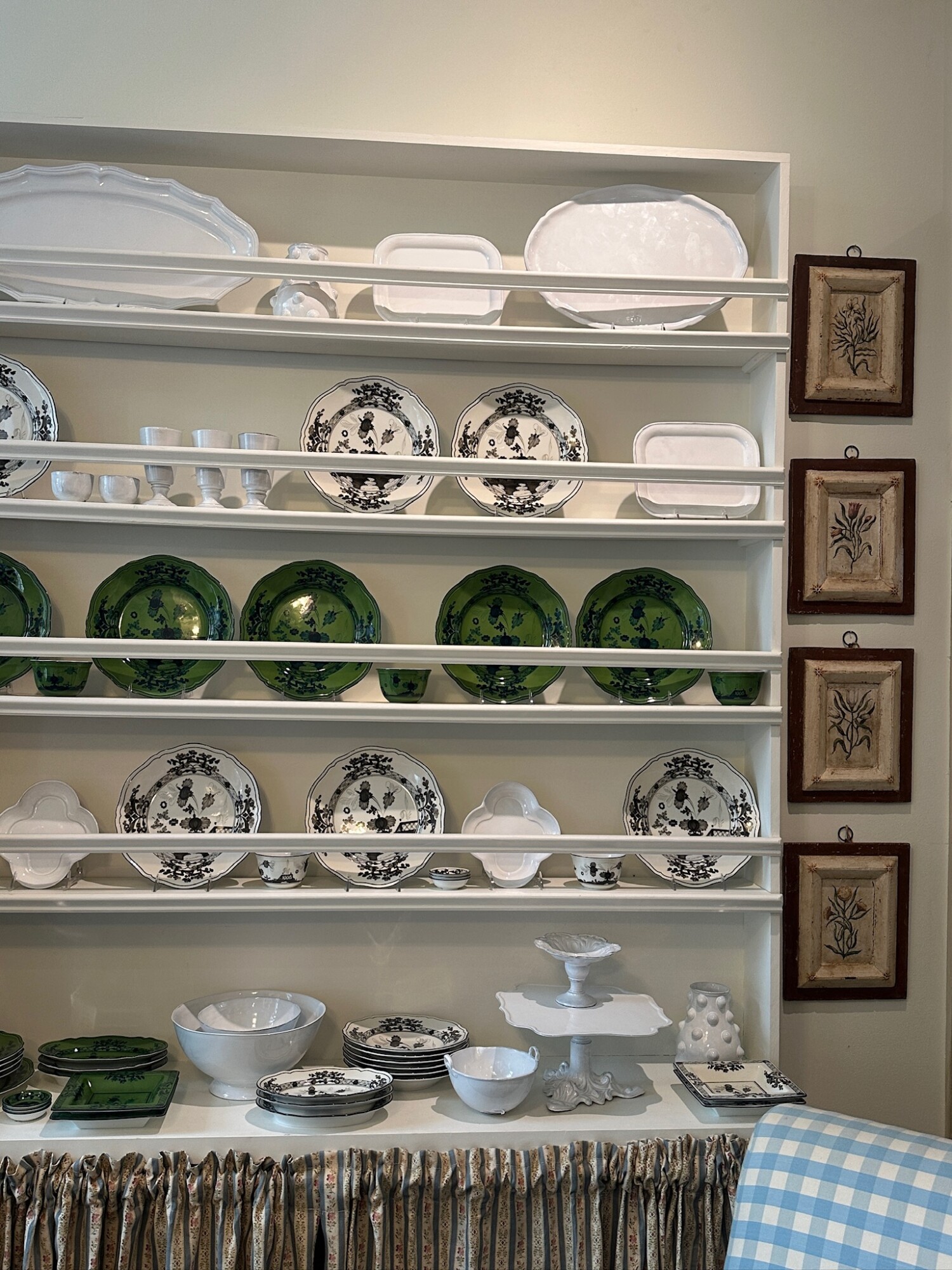 A charming open kitchen hutch displaying a curated collection of dishes and serveware. Black-and-white floral china is mixed with vibrant green plates and simple white platters. Below, cake stands and bowls are styled on a countertop skirted with a gathered floral fabric. To the right, a column of four framed botanical prints adds a vintage touch, while a blue gingham armchair peeks into the frame.