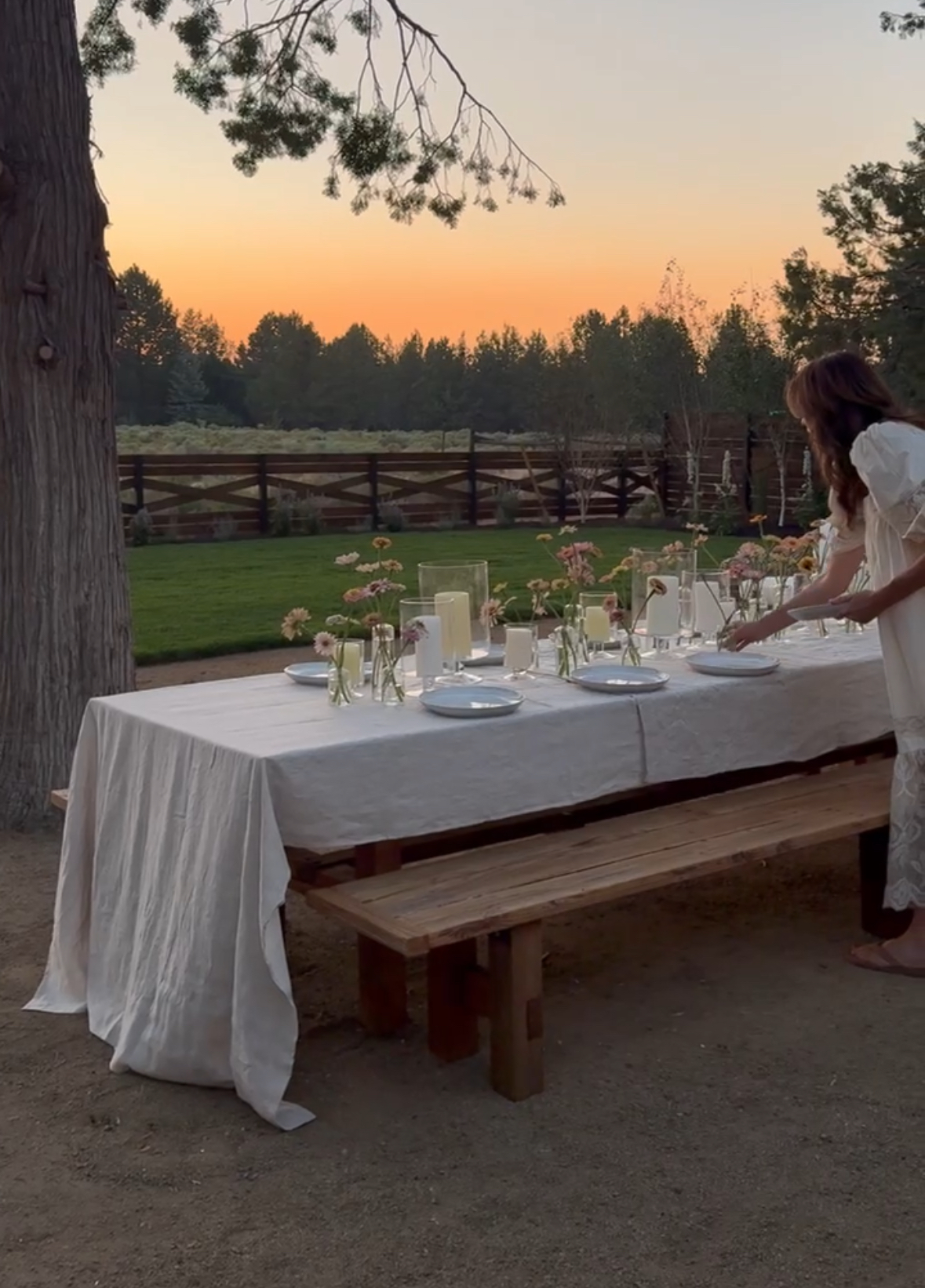 A woman sets a rustic outdoor dining table draped in a linen tablecloth, surrounded by simple wildflowers and candlelight, with a glowing sunset and wooden fence in the background—dreamy summer evening vibes on the ranch.