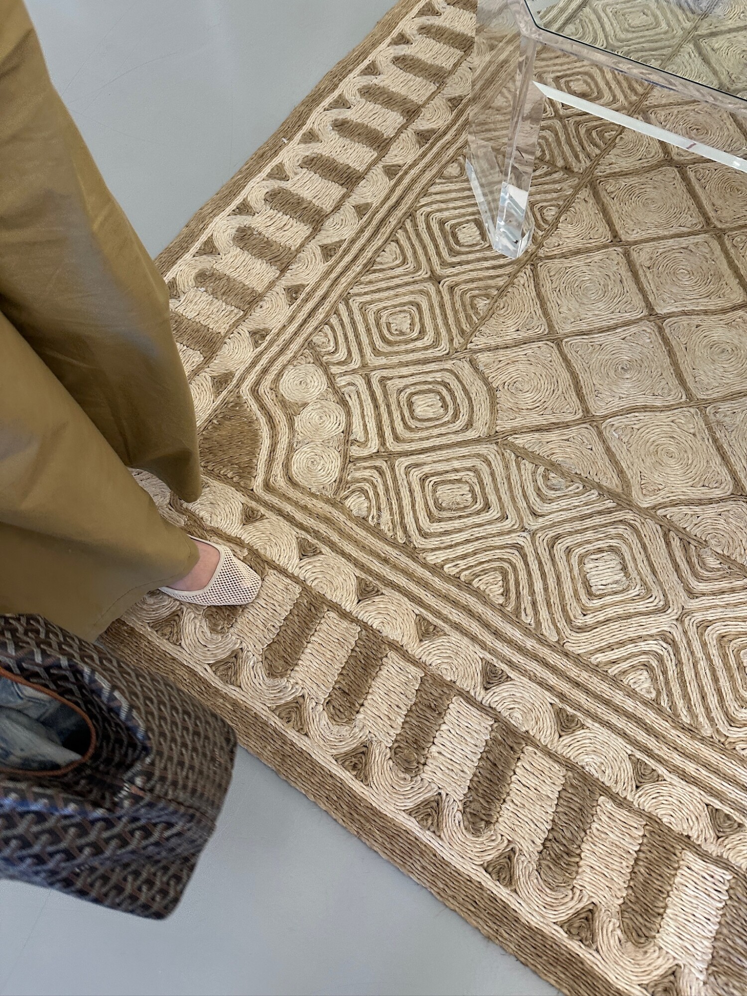 Close-up of a textured natural fiber rug with an intricate geometric pattern in soft tan tones. A woman stands partially in frame wearing wide-leg olive trousers and cream woven flats, holding a dark woven handbag. A lucite coffee table sits atop the rug, creating a modern contrast to the earthy textures below.