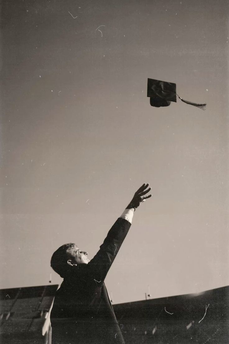 A graduate wearing a cap and gown throws their graduation cap into the air during a ceremony, captured in black and white with a nostalgic, vintage feel. The cap is mid-air against a clear sky, symbolizing achievement and new beginnings.