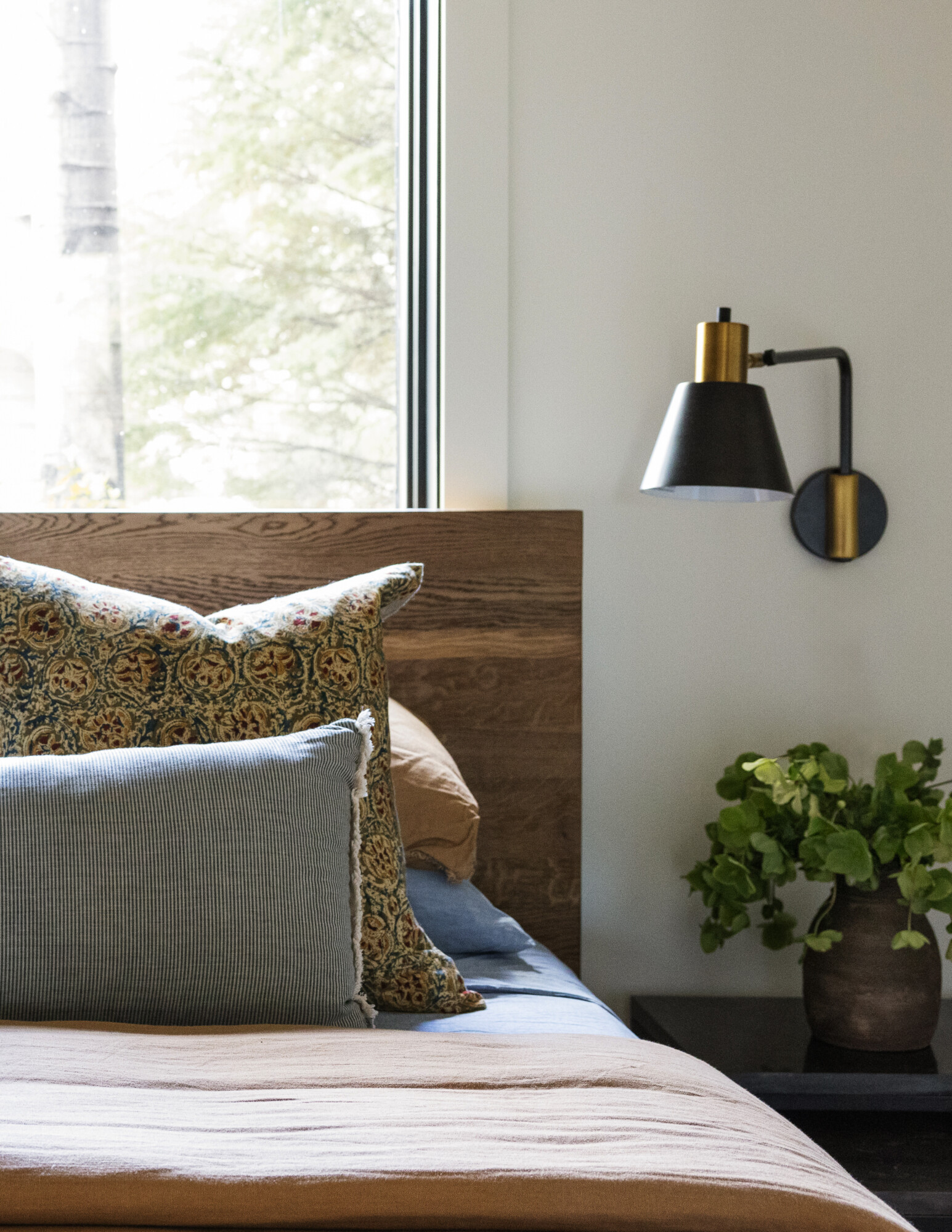 Stylish neutral-toned bed with a woven cane headboard and layered bedding in ivory and charcoal. The standout elements are a pair of large cream pillows with bold navy floral block print. Black accent pillows and a chunky knit throw add cozy texture, with a ceramic lamp and greenery on the nightstand.
