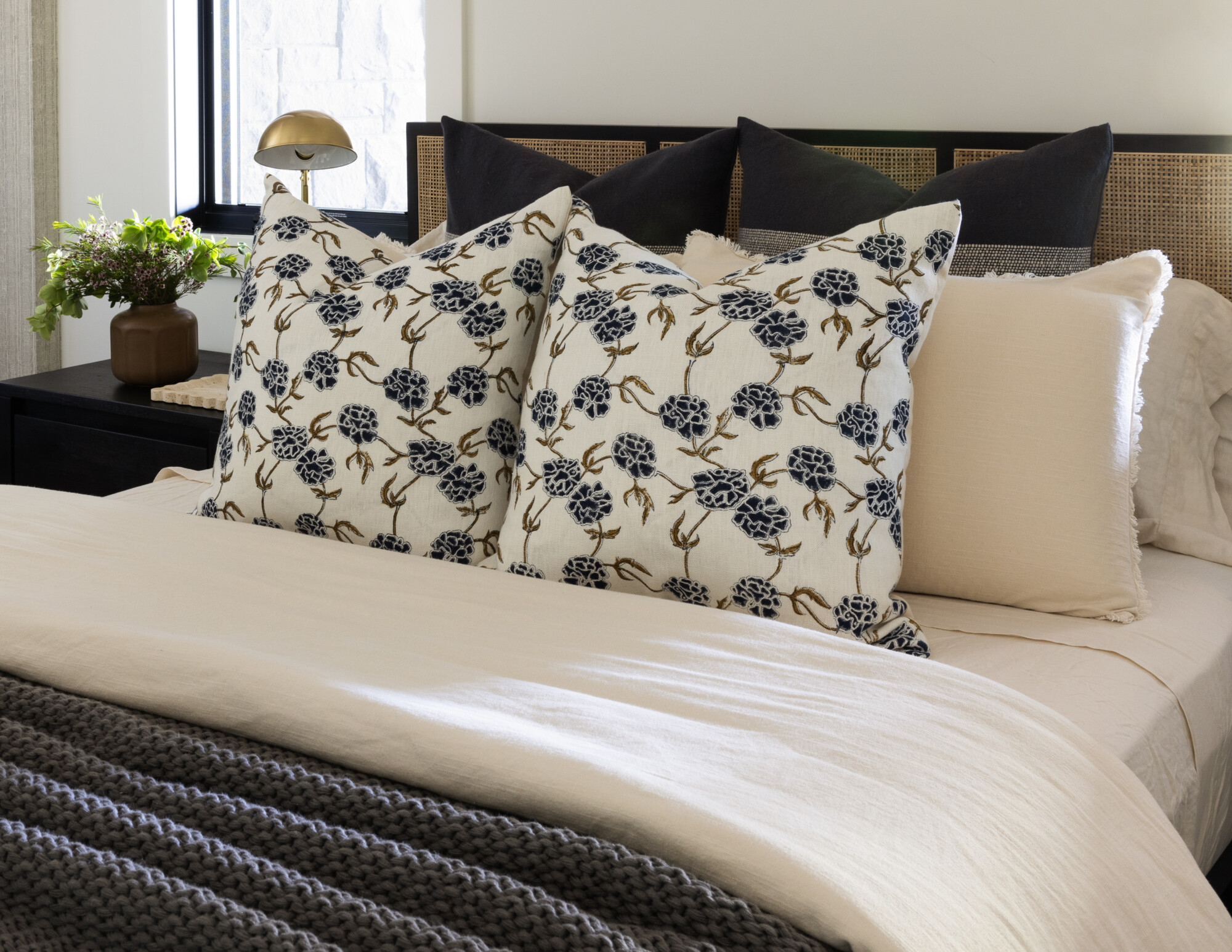 Close-up of a cozy modern bed with layered earthy bedding in warm camel and slate blue tones, accented by a wood headboard. The pillows feature a mix of floral tapestry and soft pinstripe fabrics. A black and brass wall sconce and a leafy plant on a dark nightstand add contrast and charm.