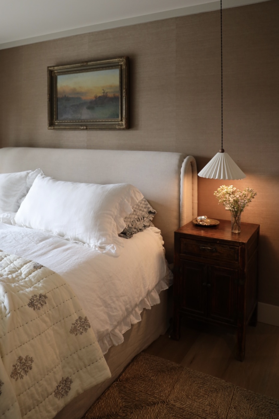 A cozy bedroom corner featuring a light upholstered bed with crisp white bedding and a quilted blanket, beside a vintage wooden nightstand topped with a small glass vase of flowers and a pleated pendant light. A moody landscape painting hangs above.