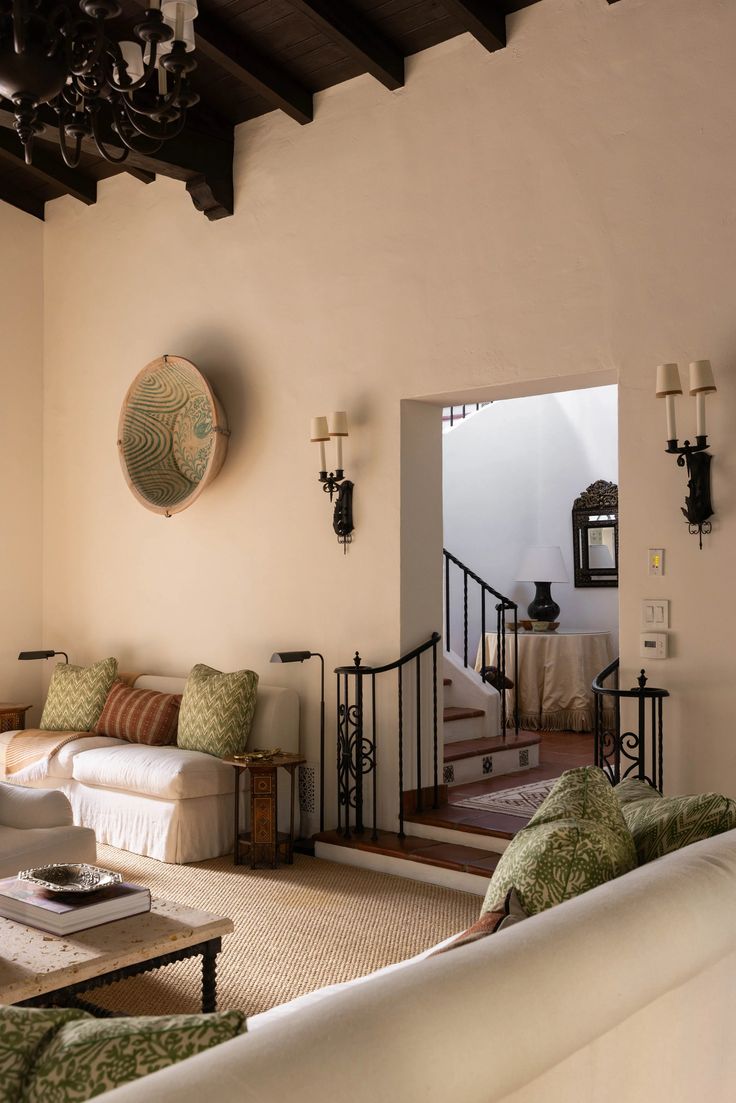 Warm and inviting Spanish-style living room with white stucco walls, dark wood ceiling beams, wrought iron railings, and soft cream slipcovered sofas accented with green and terracotta pillows. A large basket hangs as art, and layered textures like a jute rug and ornate side tables add cozy, collected charm.