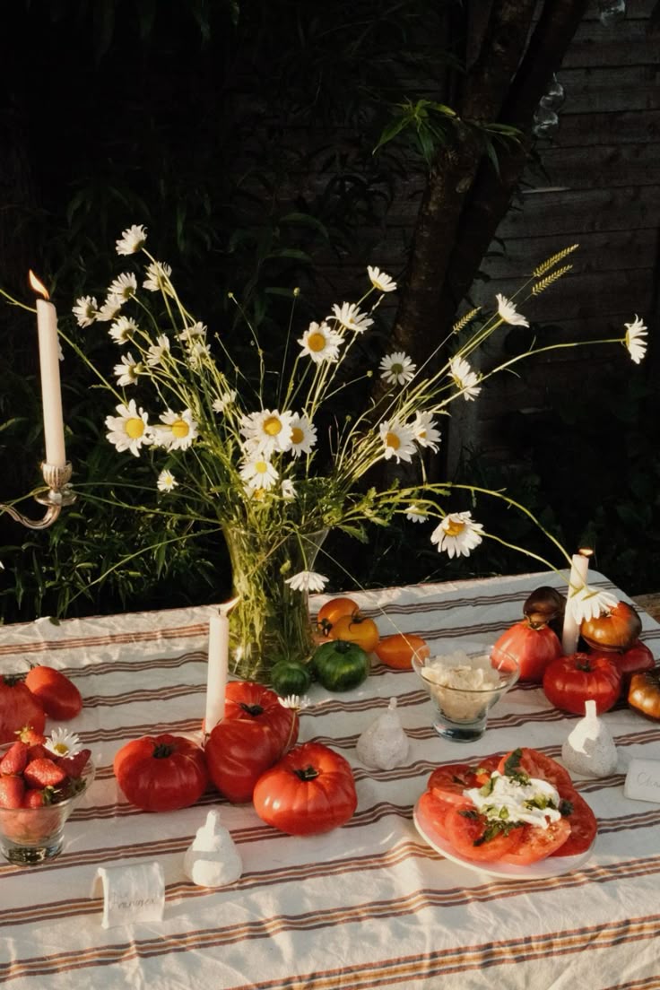 Outdoor summer tablescape featuring a striped tablecloth, taper candles, and a centerpiece of wild daisies in a glass vase. Heirloom tomatoes, fresh strawberries, and a tomato burrata salad are styled across the table in a romantic, rustic setting.