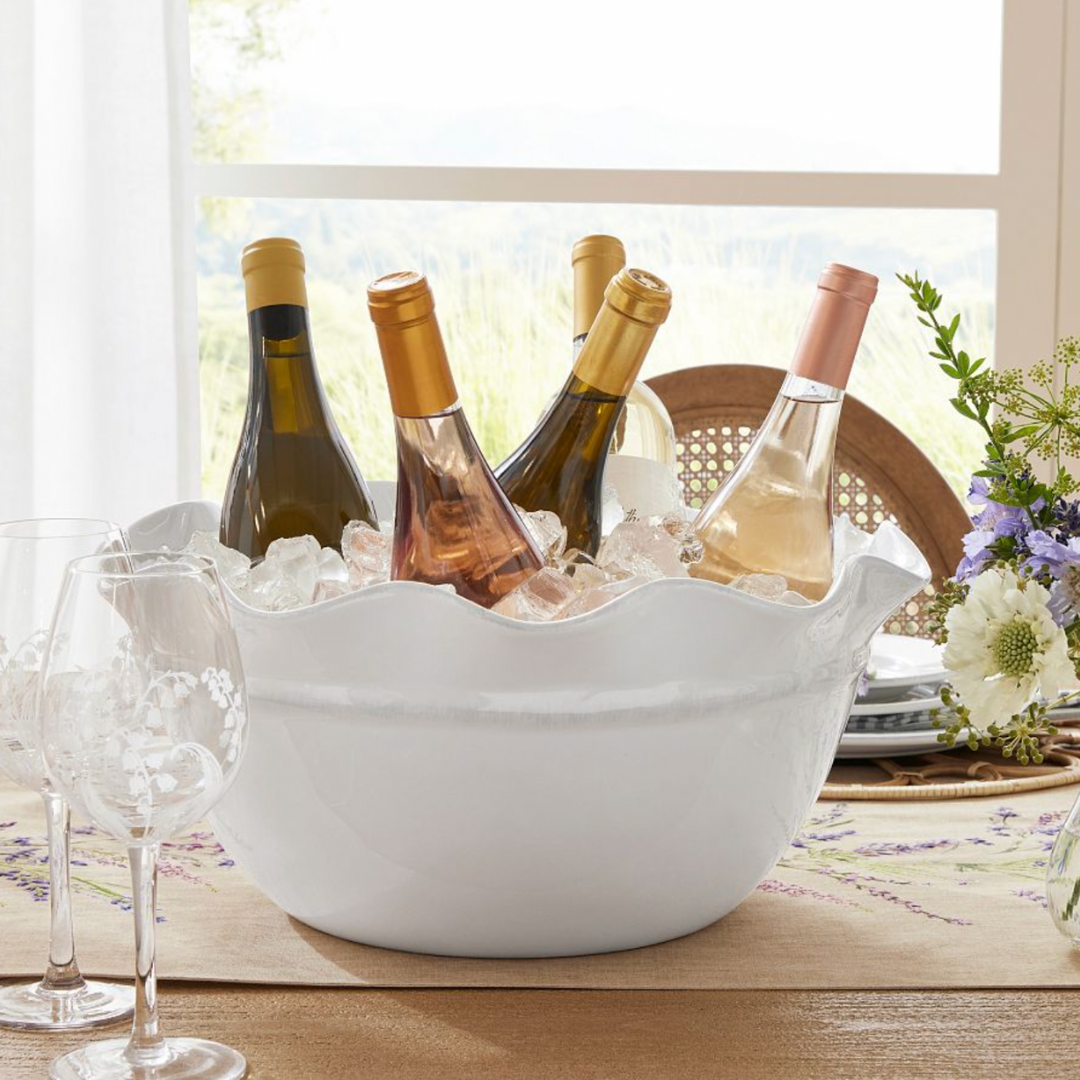 White ceramic scalloped party bucket filled with ice and assorted wine bottles, placed on a summer tablescape with etched wine glasses, fresh floral centerpiece, and natural light streaming in through the window.