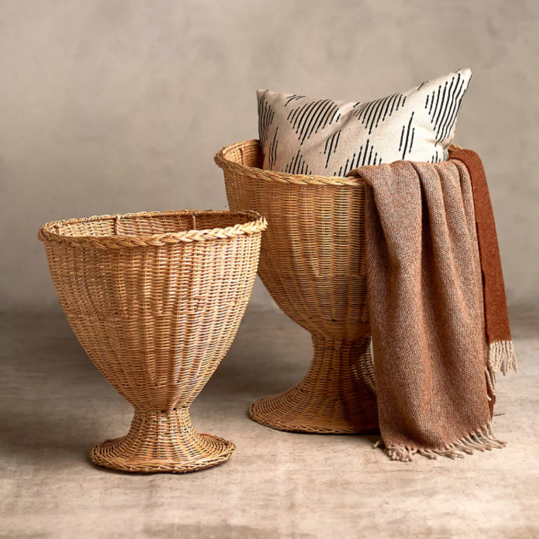 Two pedestal-style rattan urn baskets in a neutral setting, one styled with a modern patterned pillow and a rust-toned woven throw blanket draped casually over the side.