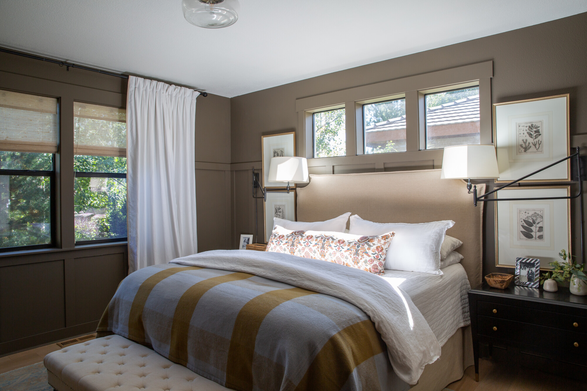 Cozy and stylish bedroom with deep taupe walls, a beige upholstered headboard, and layered white bedding. The bed features a striped mustard and gray blanket and a floral lumbar pillow. Natural light streams through wood-trimmed windows with woven shades and white curtains. Framed botanical prints, a tufted bench, and black nightstands with brass accents complete the warm, tailored look.
