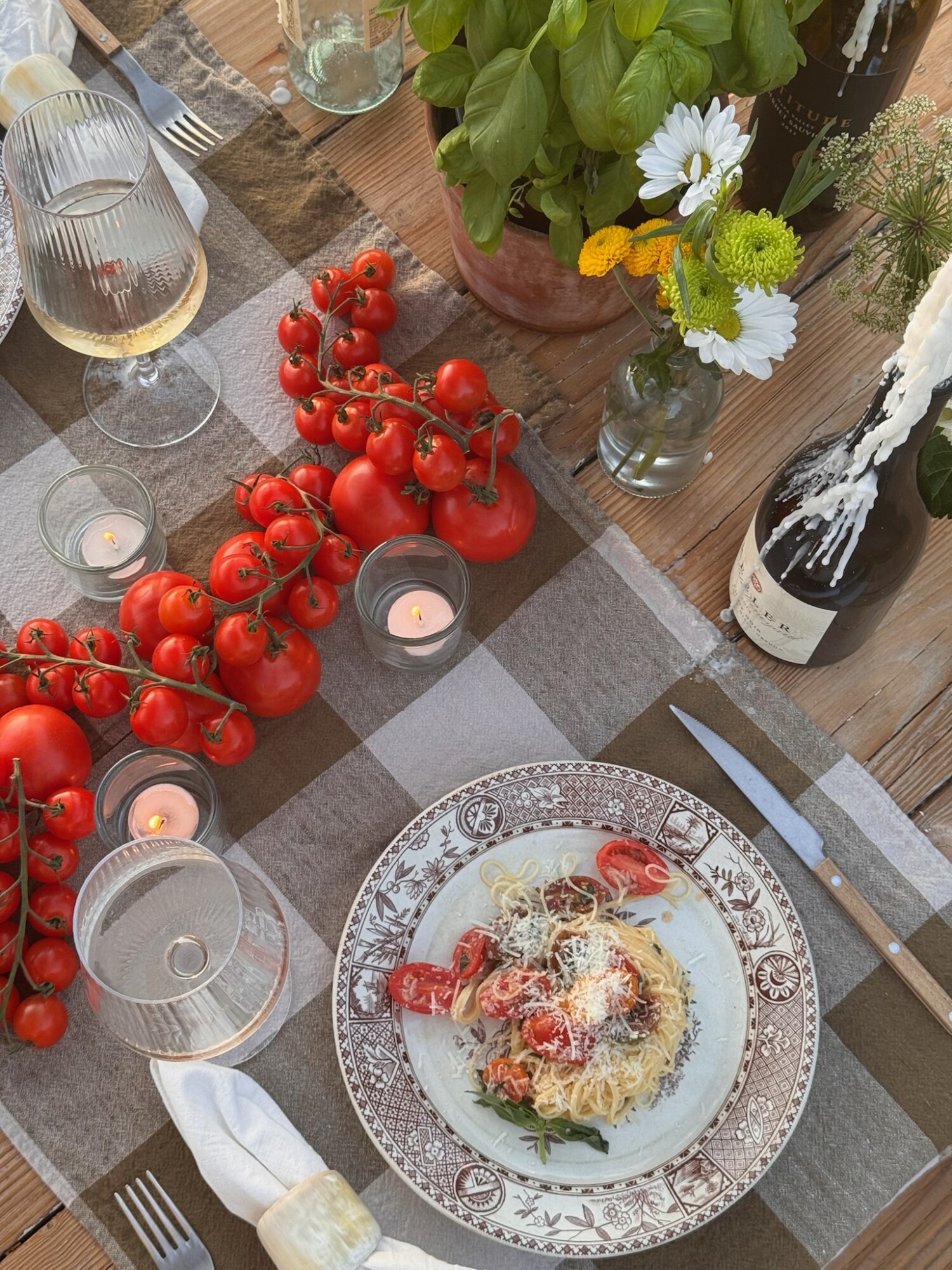 A romantic outdoor dinner setting on a wooden table with a brown gingham tablecloth, featuring a plate of summer garden pasta topped with fresh tomatoes and parmesan, surrounded by wine glasses, votive candles, a pot of fresh basil, small vases of wildflowers, and bunches of tomatoes used as a centerpiece.
