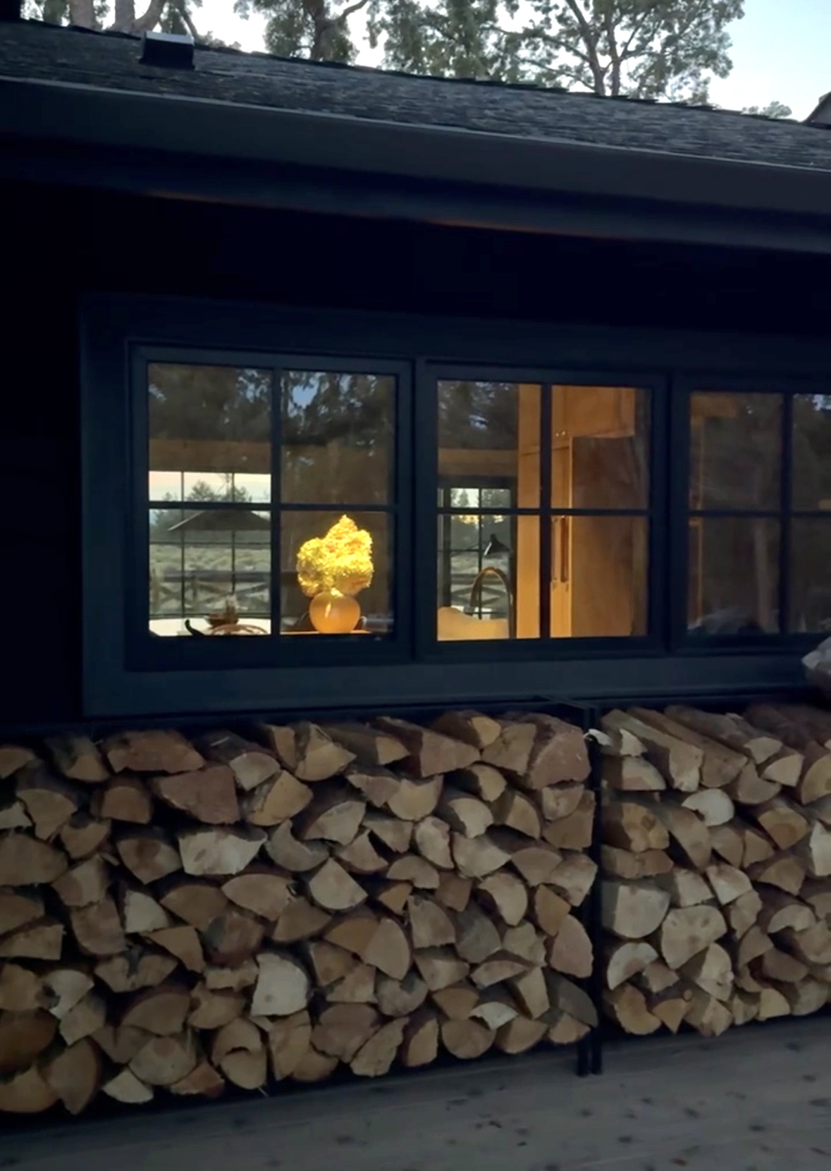 Evening view of stacked firewood beneath a dark-framed kitchen window at Tumalo Ranch. A warm glow from a lit lamp and a yellow floral arrangement is visible through the window, creating a cozy, inviting contrast against the black exterior siding.