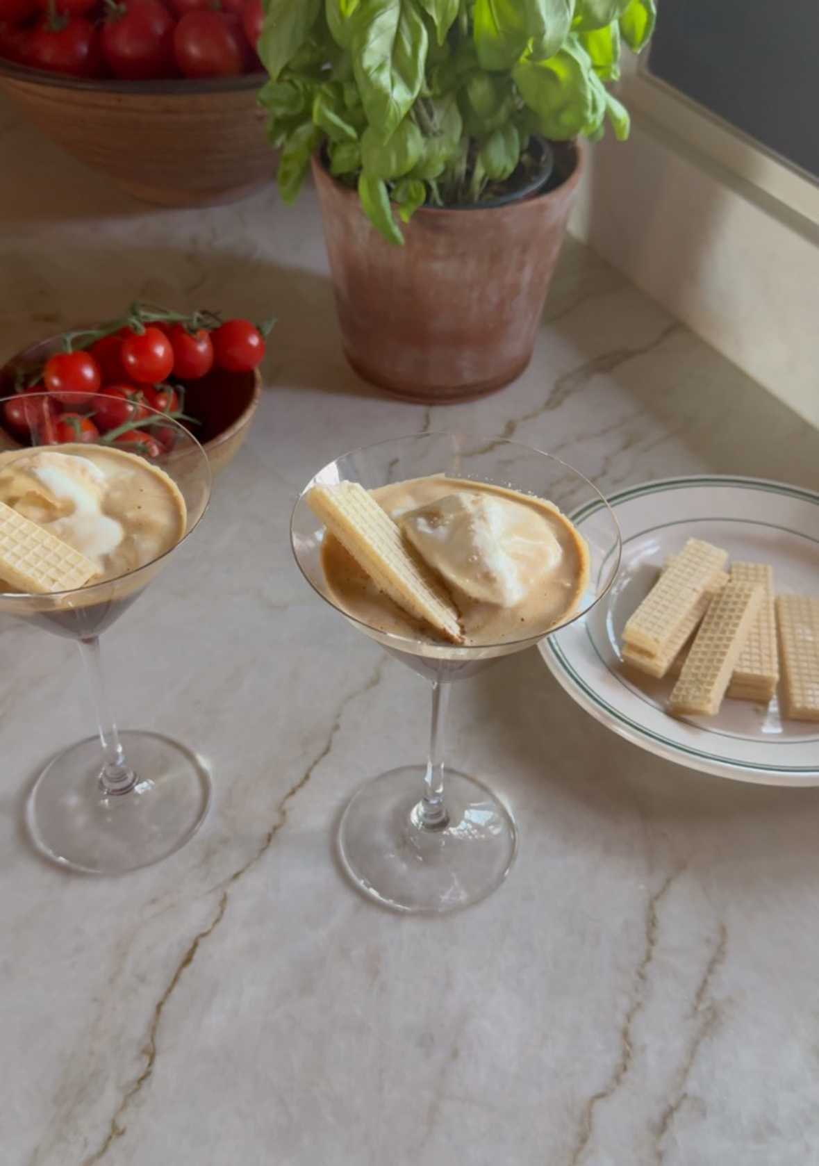 Two boozy affogato desserts served in martini glasses, each topped with vanilla ice cream and a vanilla wafer cookie, set on a marble countertop with a bowl of cherry tomatoes, a potted basil plant, and a plate of extra wafers in the background.
