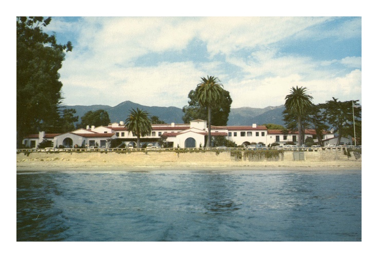 Vintage photo of the historic Four Seasons Biltmore Santa Barbara as seen from the water, with a classic white stucco and red tile roof exterior, framed by palm trees and mountains in the background under a partly cloudy sky.