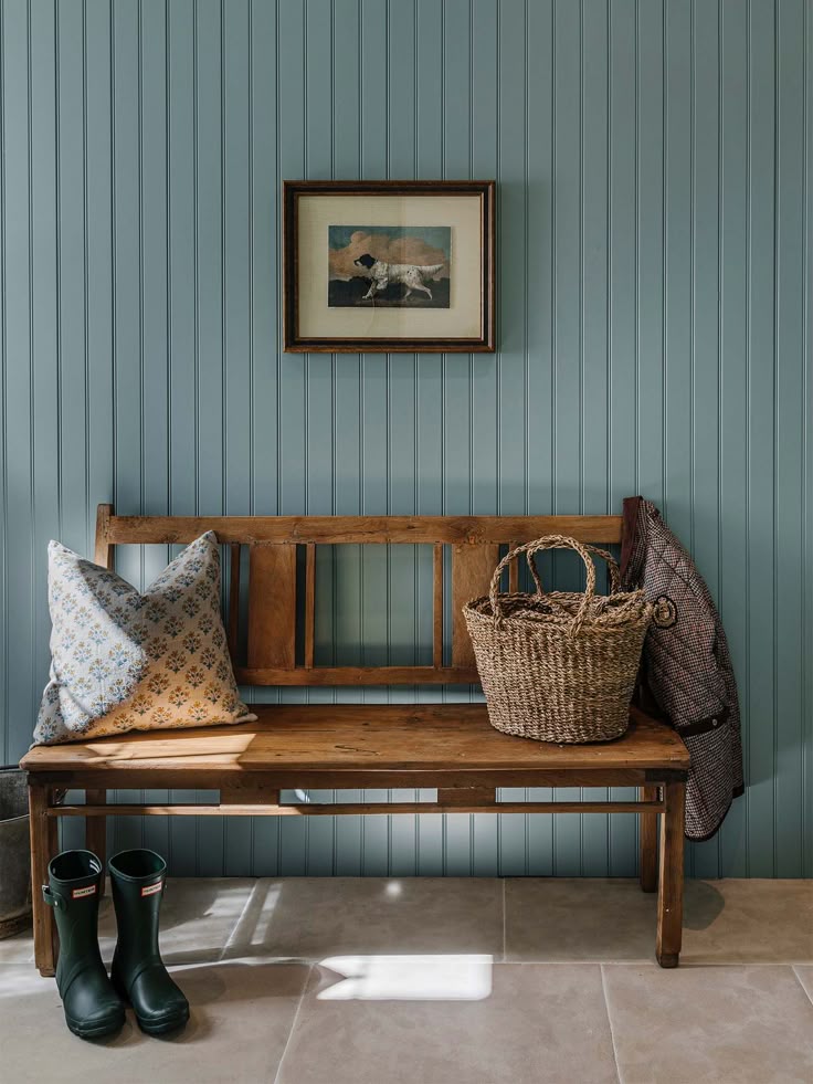 A cozy entryway featuring a wood bench set against vertical blue paneling. On the bench rests a patterned pillow, a woven basket tote, and a houndstooth coat draped on the side. Below, a pair of green Hunter rain boots sits neatly on a stone tile floor. A framed painting of a hunting dog hangs above, completing the rustic, lived-in charm.