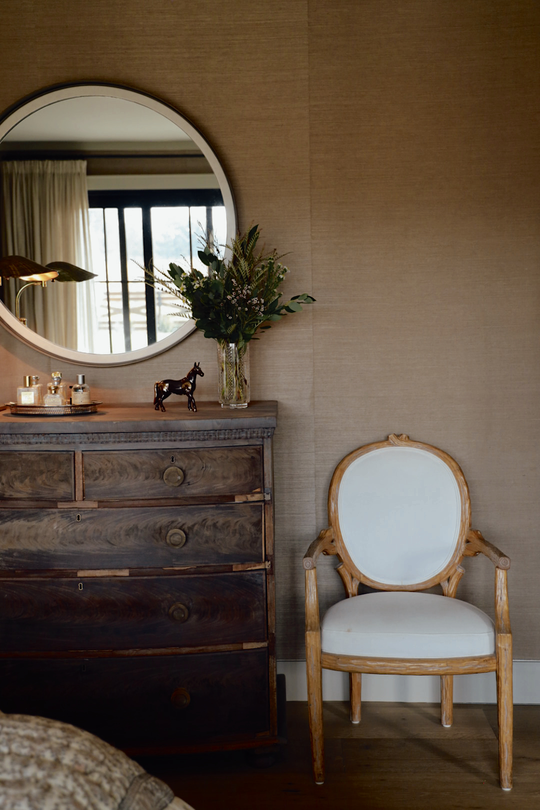Warm and moody bedroom vignette featuring a vintage wood dresser with a round mirror above, a rustic Louis-style chair with white upholstery, and layered styling with perfume bottles, a horse figurine, and fresh greenery in a glass vase.