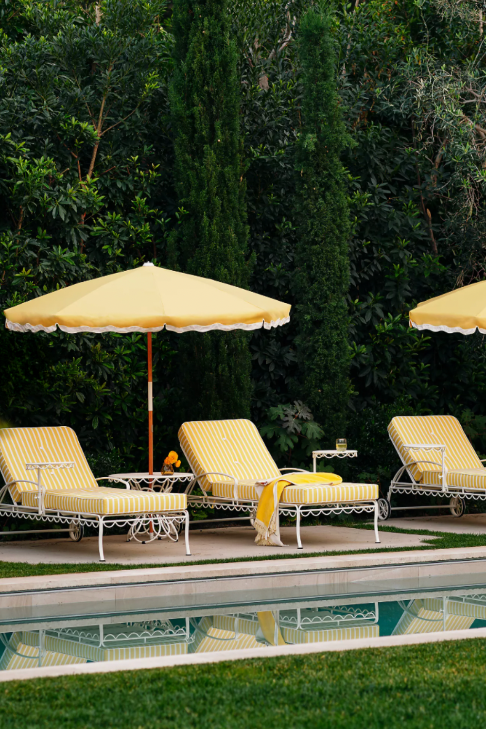 Poolside setup featuring three white metal lounge chairs with yellow and white striped cushions, matching yellow scalloped umbrellas, and small white side tables. Surrounded by lush greenery and tall trees, with the scene reflected in the clear swimming pool in the foreground.