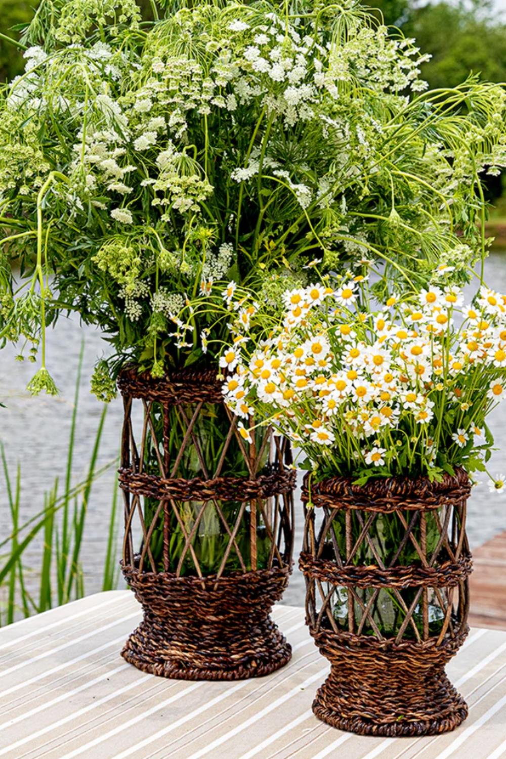 Two woven rattan hurricane candle holders repurposed as vases, filled with lush white wildflowers and chamomile, sitting on a striped outdoor table near a serene lakeside setting.