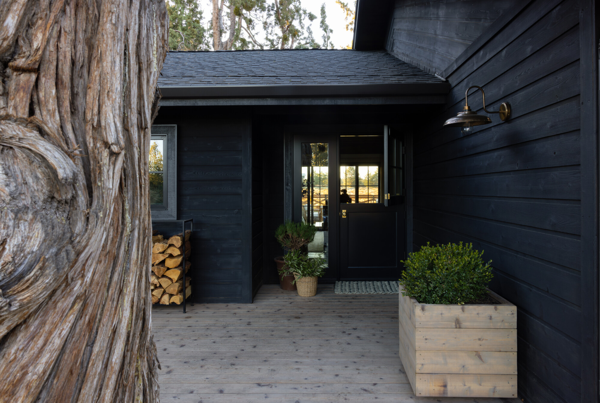 Close-up of a dark Dutch front door framed by vertical wood siding, all painted in a matte black finish. The door features a brass knob and divided glass window. A pair of potted plants—one in terracotta and the other in a woven basket—sits next to the door, adding organic texture. A woven doormat and wooden planter with boxwood complete the cozy and inviting entrance setup.