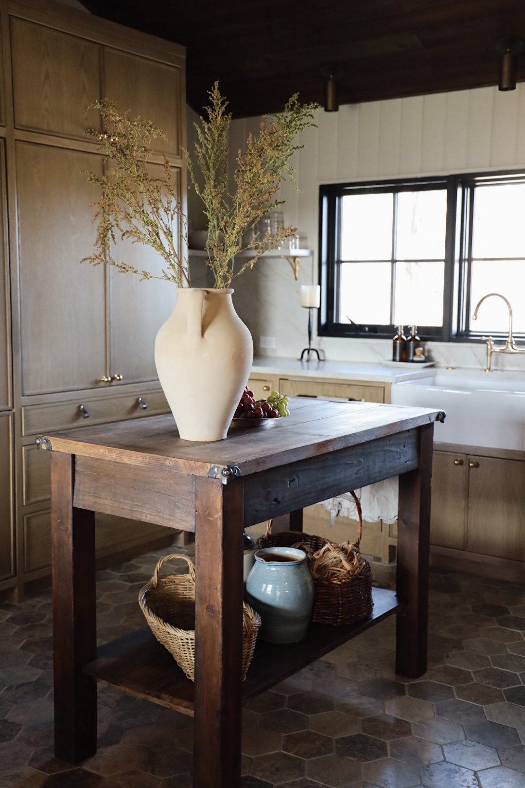 Rustic kitchen with a freestanding wooden island table topped with a sculptural cream ceramic vase holding wild branches. Below, woven baskets and a glazed pottery jug sit on the lower shelf, surrounded by natural-toned cabinetry, black-trimmed windows, and a farmhouse sink.
