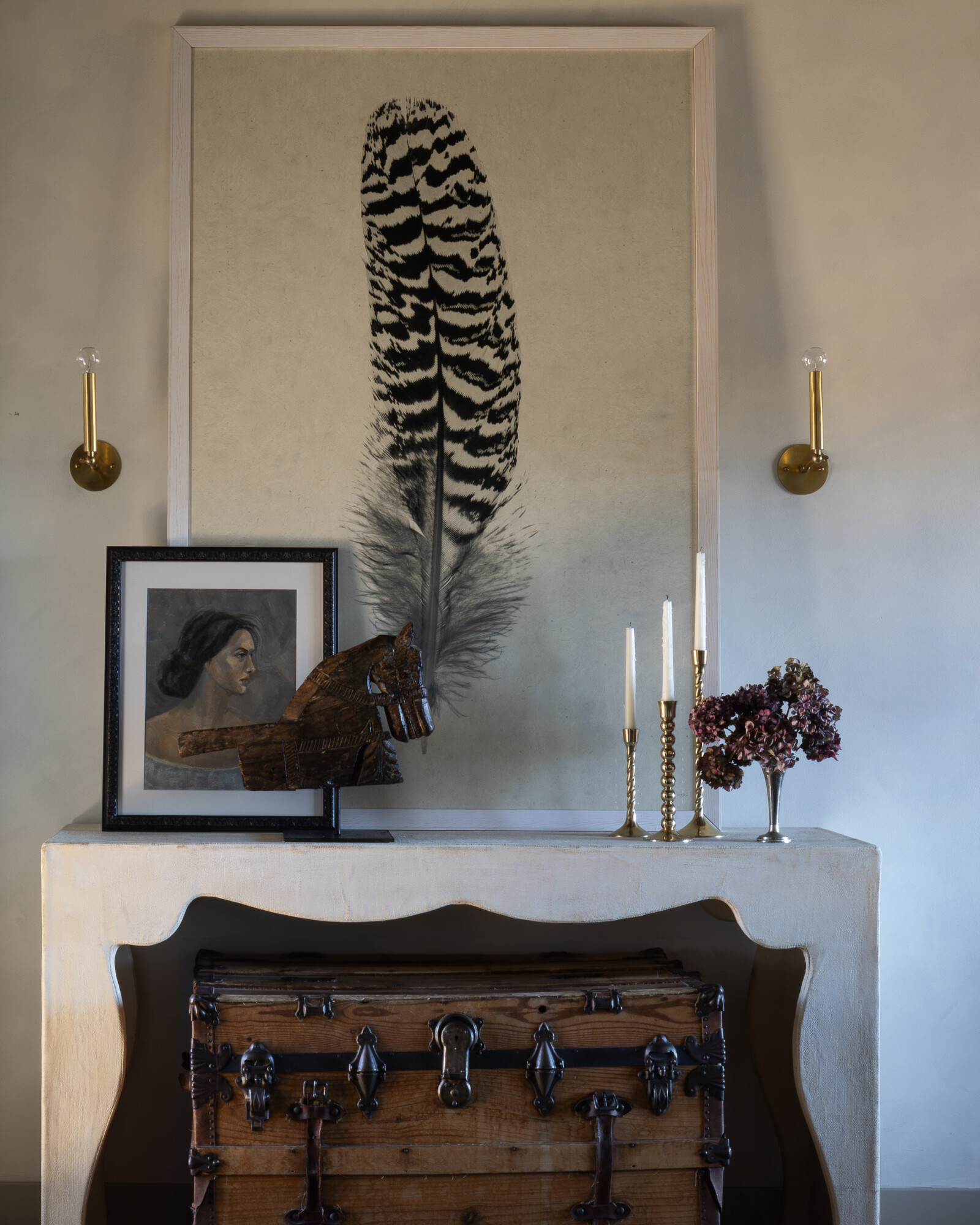 Styled entry console with vintage charm, featuring a large feather artwork, brass candlesticks, a portrait painting, wooden horse sculpture, and a weathered antique trunk underneath.
